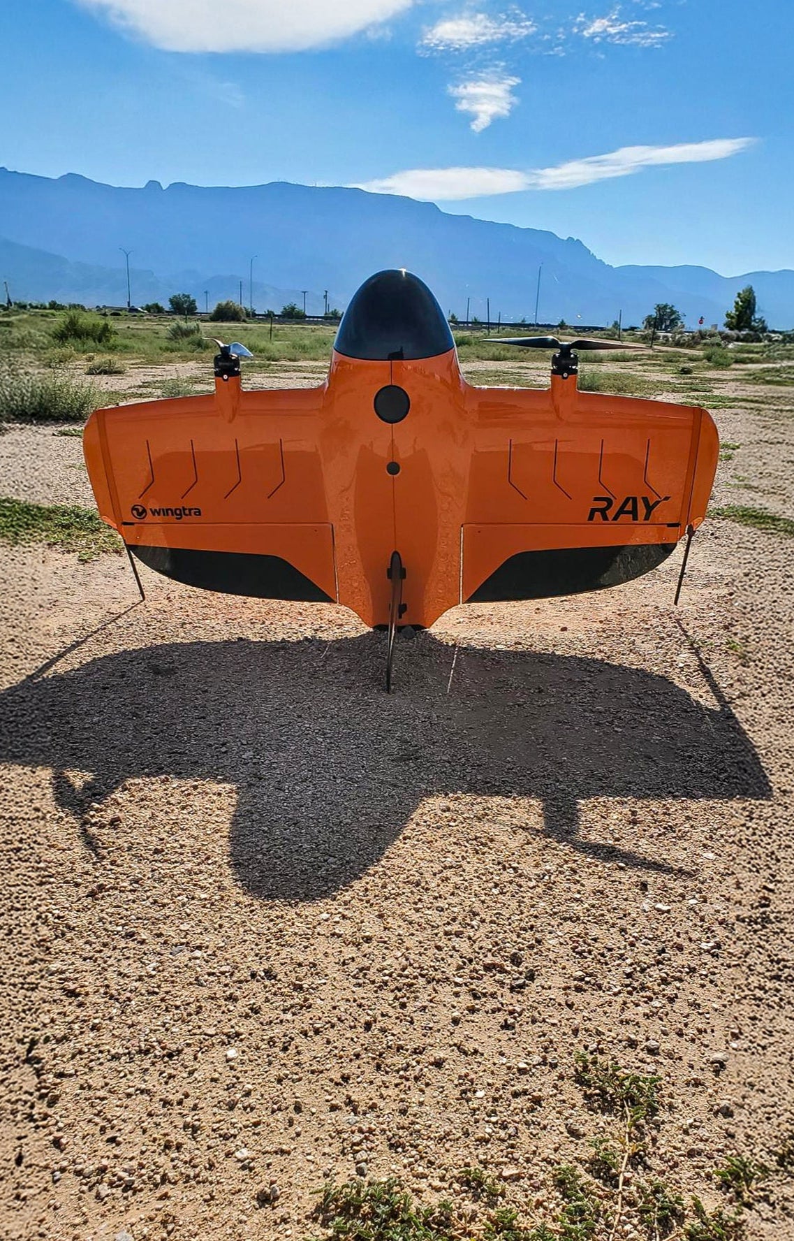 Orange and black drone with 'Ray' branding on a dirt field with mountains in the background