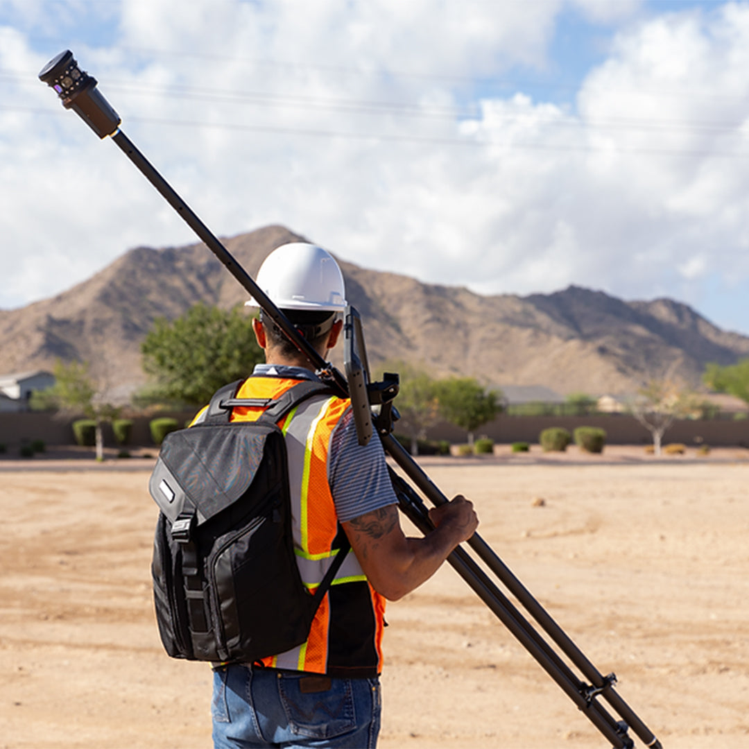 Land Surveyor in safety gear with a Grade Control Products Field Backpack and pole in an open area with mountains in the background