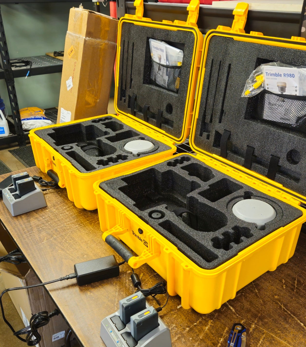 Two yellow hard protective cases opened on a wooden workbench, showing black foam inserts and accessories including gray electronic devices and a power adapter.
