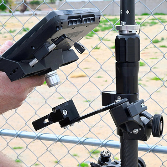 Hand holding a TSC7 with a mount attached to a tripod against a chain-link fence background