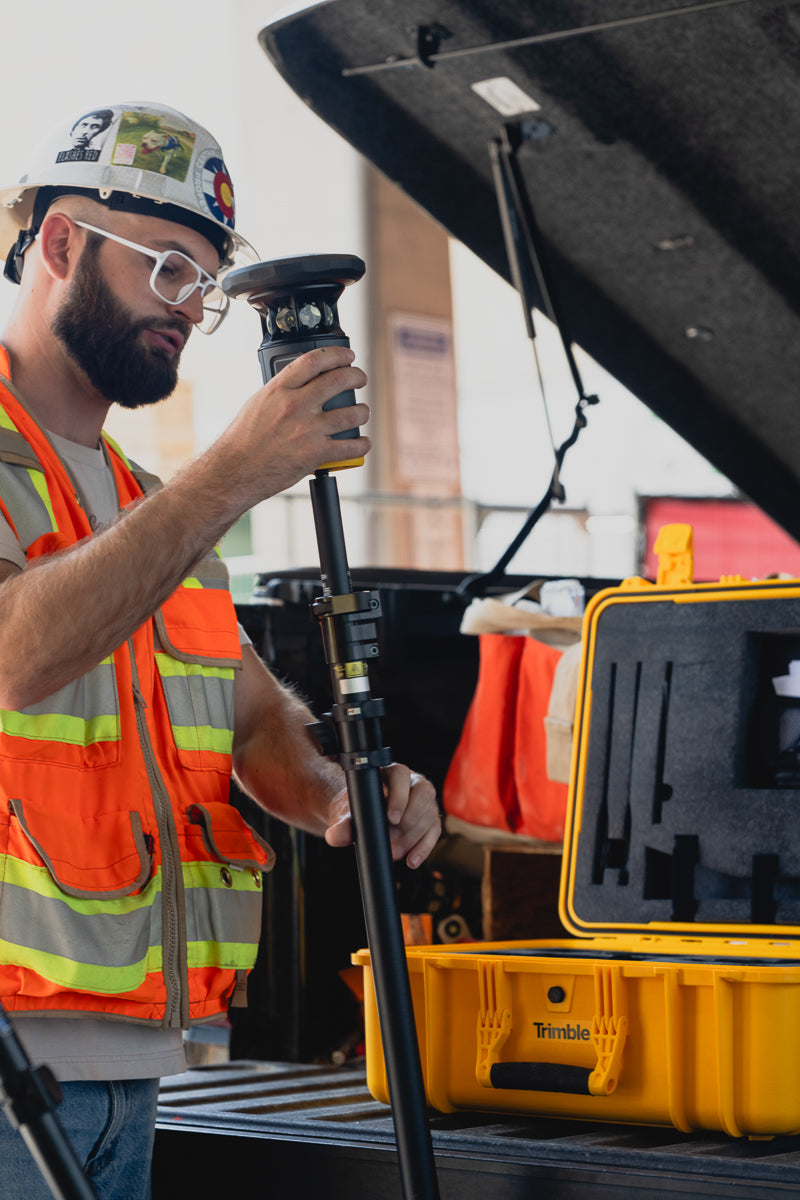 A construction worker wearing a hard hat, safety glasses, and an orange high-visibility vest operates a Trimble ST30 device mounted on a pole next to an open yellow hard case labeled 'Trimble' in a work truck bed.