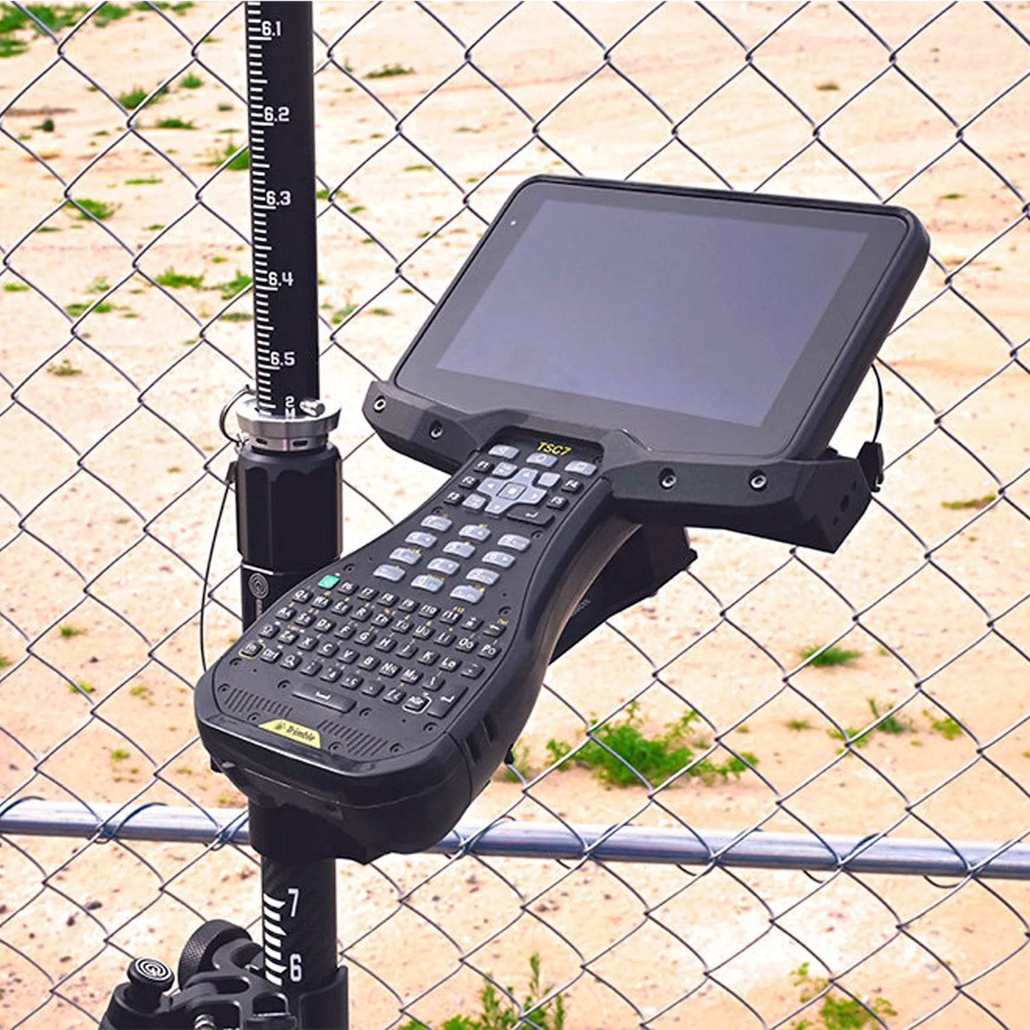 Trimble TSC7 mounted on a pole via bracket against a chain-link fence.