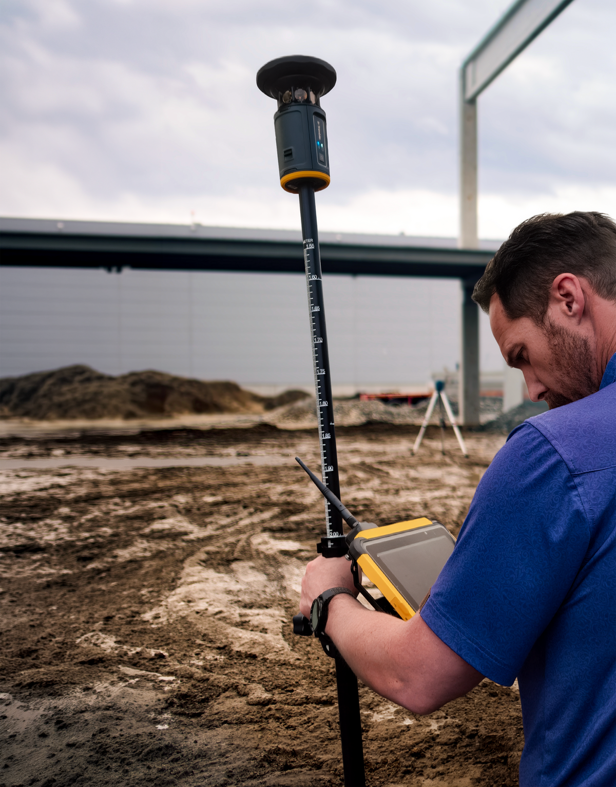 A surveyor wearing a blue shirt operates a Trimble ST30 Smart Target on a muddy construction site under an overcast sky.